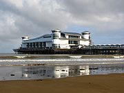 A walkway supported by metal legs arising from the sea, at the end of which is a large a white building with a wave-shaped roof and corner turrets.