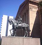 Equestrian statue in dark grey metal of George V in military dress uniform on a plinth of red granite outside a Classical building of red sandstone