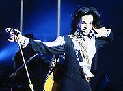 Waist high portrait of thirty year old male, wearing longish hair and short beard, and a black jumpsuit over one shoulder, right hand extended to hold microphone stand. Shirt has unnamed 