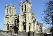 A stone built Victorian Gothic building with two square towers and a central arched entrance underneath a circular ornate window. A Victorian street lamp stands in front of the building and on the right part of a leafless tree, with blues skies behind.