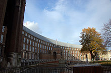 A large brick building, built in a shallow curve, with a central porch. In front of that a pool and a water fountain. Autumn trees on the right and a blue sky with some clouds above.