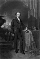 Engraved full-length portrait of a balding man standing next to a table with his left arm resting on a book and in the background a stone balustrade beyond which are trees and a building with columned portico