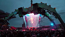 A concert stage; four large legs curve up above the stage and hold a video screen which is extended down to the band. The legs are lit up in green. The video screen has multi-coloured lights flashing on it. The audience surrounds the stage on all sides.