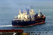 A large, almost rectangular black ship with a red hull creates a mild wake as it moves away from the shore into a very large river. Smoke rises above its decks, which are cluttered with antennas, mechanical fittings, and what appear to be self-contained rooms with multiple windows.