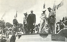 Five people on stone platform, from left: Man with mustache in military dress uniform and bushy tall black hat with chinstrap; man in military uniform with many medals and old fashioned naval officer's hat; man with white hair in suit with long dark coat and white collar; woman in Victorian dress with white hat; man with beard and wild hair in suit. Crowd and flags in background.