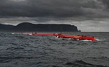 A long red tube lies in the water under dark, cloud-covered skies with black hills in the distance.