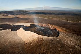 A column of white smoke rises off a simmering black-topped lava lake in the center of a large semi-circular crater. Its most recent eruptive product has left a dark channel leading off to the left, and the terrain surrounding the cone is sparse; another column of smoke and a large shield-shaped mastiff rise in the background.