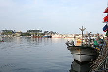 Two boats anchor in a harbor on the right. Blue skies and the sea are clear and tranquil.