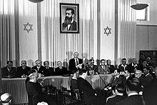 A man reads a document to a small audience assembled before him. Behind him are two elongated flags bearing the Star of David and portrait of a bearded man in his forties.