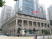 The grey dome and front gable of a granite neo-classical building, with a skyscraper in the background against a clear blue sky