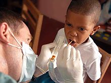 white man holding plastic tray with brown goop in it and sticking a small stick into a black boy's open mouth