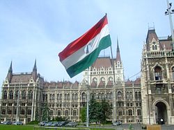 Tricolor Hungarian flag with circle cut from the middle flying atop a bent flagpole in front of a large Neogothic building