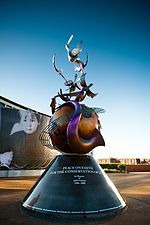  A colour photograph of a large metal monument with a conical base supporting a globe that is wrapped in contorted musical instruments. In the background is a blue sky.