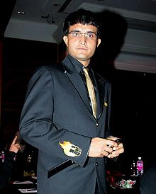 Clean-shaven man wearing glasses stares directly at the camera. He is wearing a black suit and a gold tie. The man's right arm is facing the camera. The arm has a small picture of a helmet engulfed with fire.